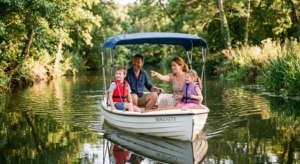 Une famille avec deux enfants naviguant paisiblement sur une petite rivière calme à bord d'un bateau électrique blanc, sous un soleil doux de fin d'après-midi.