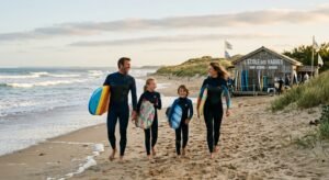 Une famille avec enfants souriants portant des planches de surf sur une plage de Vendée, devant une école de surf conviviale en bois sous un ciel ensoleillé.
