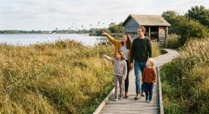 Une famille avec deux jeunes enfants marchant tranquillement sur un sentier en bois au bord d'un étang dans une réserve naturelle, avec un observatoire en bois en arrière-plan sous un soleil doux.