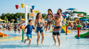 Une famille souriante profite d'une zone de jeux aquatiques colorée et ensoleillée dans un parc aquatique, avec des jets d'eau et des toboggans en arrière-plan.